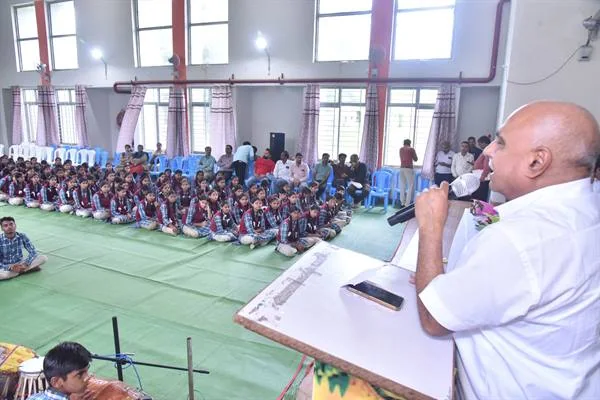 विद्यालय के सभागार में मंच से माइक पर बोलते व्यक्ति के सामने फर्श पर बैठे विद्यार्थी