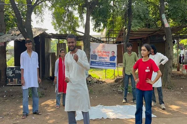 खुले स्थान पर नुक्कड़ नाटक प्रस्तुत करते युवा कलाकार, सामने सफेद कुर्ते में संवाद बोलता कलाकार और लाल टी-शर्ट में खड़ी युवती