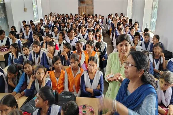 कक्षा में बैठी बड़ी संख्या में छात्राओं के बीच सामने खड़ी दो महिलाएं बातचीत करती हुईं