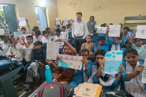 कक्षा में बैठे छात्र-छात्राएं अपने बनाए चित्र दिखा रहे हैं और शिक्षक उनके बीच खड़े हैं