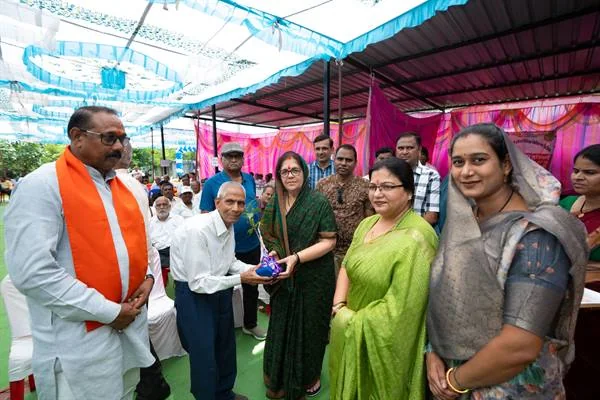 समारोह में एक बुजुर्ग व्यक्ति को पैकेट देती महिला, पास खड़े अन्य अतिथि और उपस्थित नागरिक