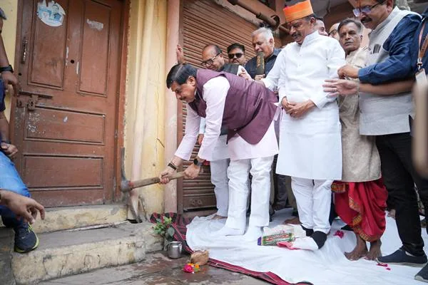 मुख्यमंत्री डॉ. मोहन यादव एक पुराने मकान के लकड़ी के दरवाजे के पास झुककर गेती चला रहे हैं; पास में सफेद चादर पर पूजा सामग्री, लोटा और फूल रखे हैं और आसपास सफेद कुर्ता-पायजामा पहने लोग तथा अधिकारी खड़े हैं।